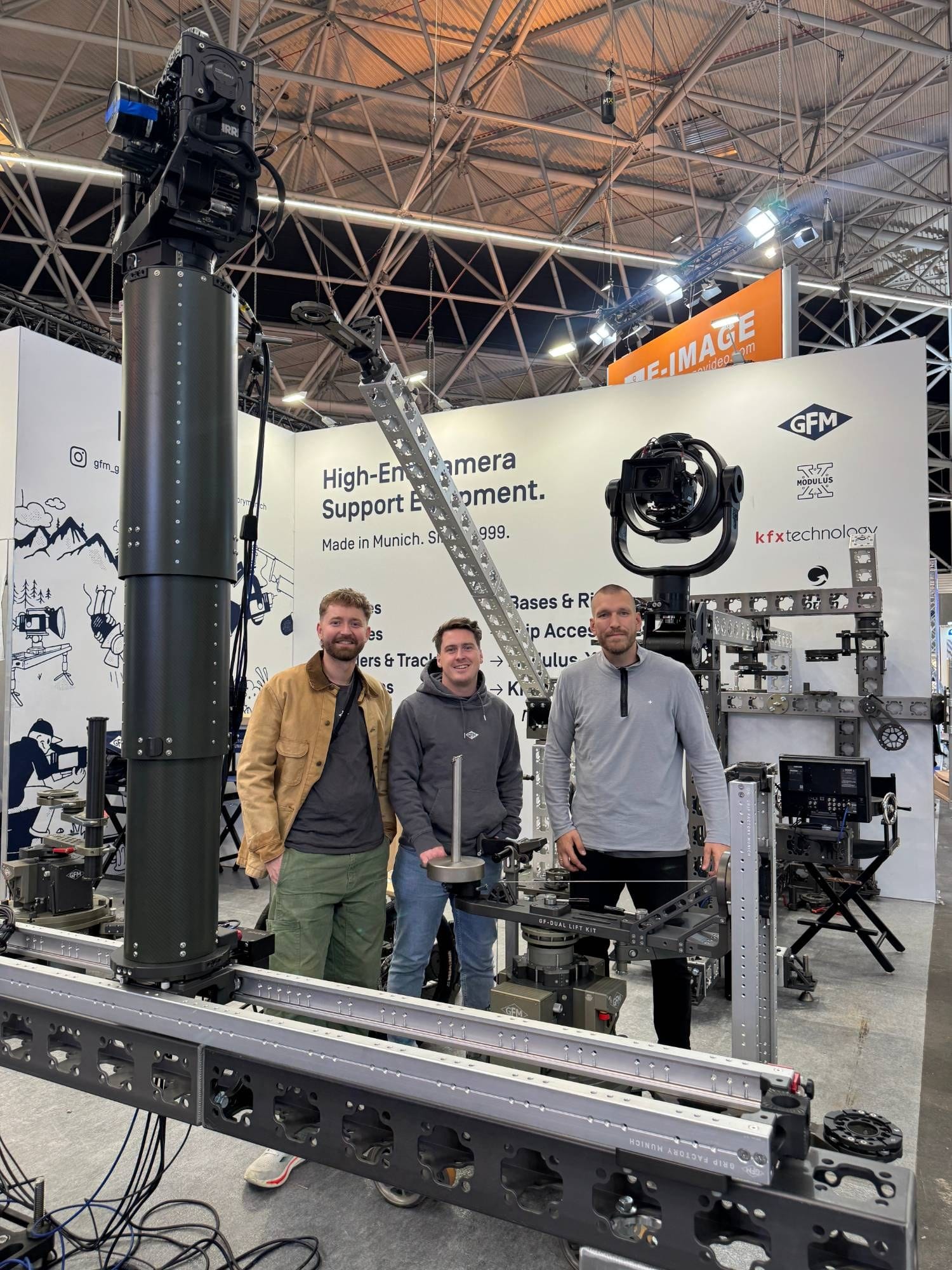 Three members of the Grip Factory Munich team on the GFM stand at IBC 2026, between a carbon camera column carrying an ARRI camera, a Modulus-X® jib, a remote head and the GF-Dual Lift Kit